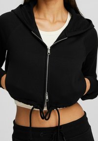 Felpa nera corta con zip argentata, orlo con coulisse e cappuccio. Presenta polsini a coste e uno strato interno cremoso a contrasto.