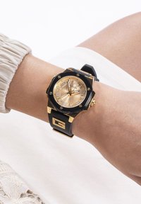 Schwarze Gummiband-Uhr mit einem goldfarbenen achteckigen Gehäuse, goldenem Zifferblatt und schwarzen Akzenten. Verfügt über große Stundenmarker und ein Markenlogo.