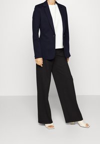 Blazer navy con solapas de muesca, dos bolsillos delanteros y cierre de un solo botón, combinado con pantalones de pierna ancha negros y sandalias blancas.