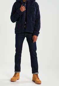 Mężczyzna w granatowej polarowej kurtce, ciemnych dżinsach i beżowych sznurowanych butach, stojący na tle jednolitego jasnego tła.