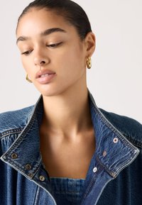 Νεαρή γυναίκα με ανακατεμένα πίσω μαλλιά, φορώντας χρυσούς κρίκους στα αυτιά, μια μπλε τζιν μπουφάν με σηκωμένο γιακά, κοιτάζει προς τα κάτω μπροστά σε ένα απλό φόντο.