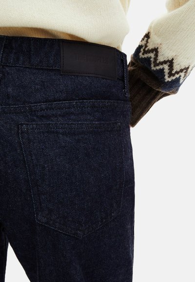 Jeans de ganga escura com um acabamento texturizado, apresentando um patch de pele preta na cintura e um design clássico de cinco bolsos.