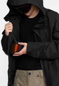 Svart vattentät jacka med huva, håller i skidglasögon med orange lins, tillsammans med beige byxor. Texturerad tyg synlig i jackan.