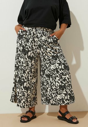 Frau trägt schwarz-weiß gepunktete, weit geschnittene Hose, schwarzes Oberteil und schwarze Sandalen mit Schnallen, steht mit Händen in den Taschen.