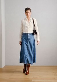 Baltas rakstains trikotāžas blūzs, augstas jostasvietas zilas džinsa svārki un brūnas augstpapēžu zābaki. Īsi mati un brūna rokassoma noslēdz apģērbu.