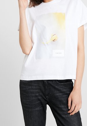Woman wearing white Calvin Klein T-shirt with a yellow flower graphic and black jeans, hand near face and relaxed pose.