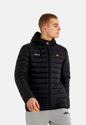 Muž v čiernej prešívanej bunde s kapucňou a zipsom s logom ellesse, pozerá sa do strany, ruky uvoľnené pri páse, na jednofarebnom pozadí.