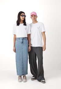 Dos personas están de pie una al lado de la otra. Una lleva una camiseta blanca corta y una falda de mezclilla azul claro; la otra lleva una camiseta blanca y pantalones a rayas.