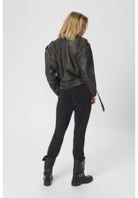 Szőke nő fekete bőrkabátban, fekete szűk nadrágban és fekete csatos csizmában, háttal áll egy egyszerű világos háttér előtt.