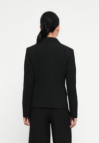 Blazer noir avec une coupe ajustée, design à un seul boutonnage, épaules structurées et tissu lisse. Comprend une fente au dos et des coutures minimales.
