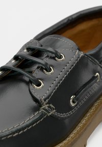 Schwarze Lederbootsschuh mit kontrastierenden weißen Nähten, runden Metall-Ösen und einer strukturierten braunen Sohle. Schnürsenkel durch die Ösen gefädelt.