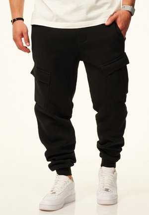 Mann trägt schwarze Cargo-Jogginghosen und weiße Sneakers, steht mit einer Hand in der Tasche vor einem schlichten Hintergrund.