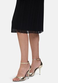 Gouden hoge haksandalen met een enkelbandje, open teen ontwerp en een glanzende afwerking; gecombineerd met een zwarte geplooide rok.