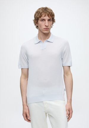 Jeune homme aux cheveux blonds bouclés portant un polo bleu clair à manches courtes et un pantalon blanc, debout devant un fond blanc uni.