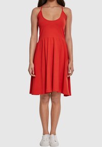 Robe sans manches rouge avec un décolleté arrondi, corsage ajusté et jupe évasée. Fabriquée en tissu lisse, arrivant à mi-cuisse.
