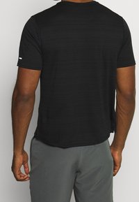 Man wearing a short-sleeve black t-shirt and gray pants, shown from the back against a plain light gray background.