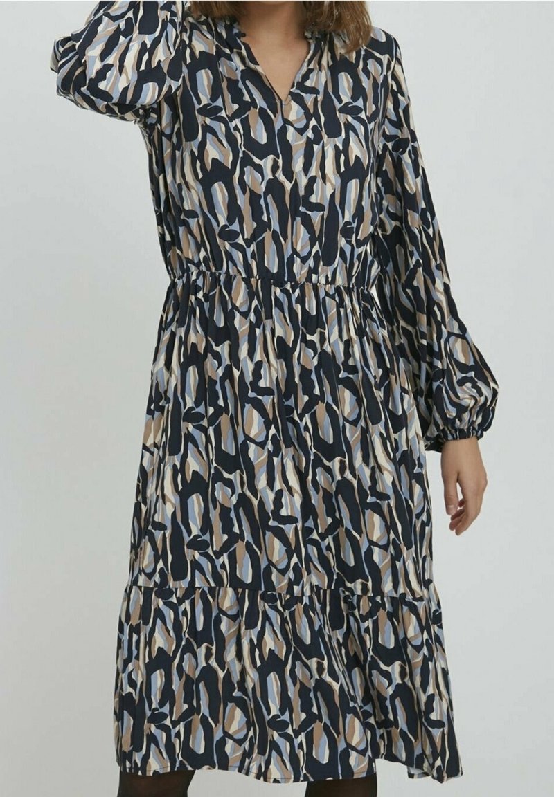 Femme portant une robe longue à manches longues, de longueur mi-mollet, avec une taille froncée et un motif abstrait en noir, beige, blanc et nuances de bleu.