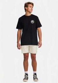 T-shirt nera in cotone con maniche corte e un logo rotondo sul petto. Abbinata a pantaloncini beige e sneakers alte nere.