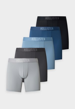 Homme musclé à la peau foncée, debout pieds nus, portant un boxer Hollister bleu clair sur un fond clair uni.
