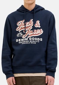 Person trägt einen marineblauen Hoodie mit dem Text "Jack & Jones Denim Goods Superior Quality Durable Wear" und einer Grafik auf der Vorderseite.