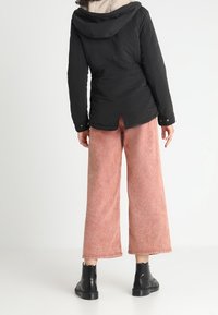 Personne debout, dos tourné, portant une veste noire à capuche, un pantalon large rose et des bottines noires sur un fond blanc.