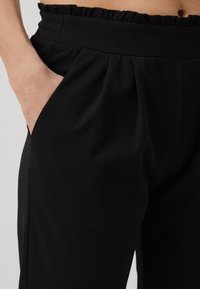 Gros plan d'une personne portant un pantalon noir taille haute avec une ceinture froncée et des poches latérales, main partiellement dans une poche.