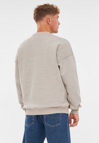 Sudadera de color beige claro con cuello redondo y hombros caídos. Presenta puños y dobladillo de canalé, combinada con jeans azules y bolsillos traseros visibles.