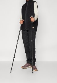 Schwarze Fleece-Weste mit Reißverschluss, kombiniert mit einem hellen Langarmshirt, schwarzen Hosen und Trekkingstöcken. Braune und graue Sportschuhe.