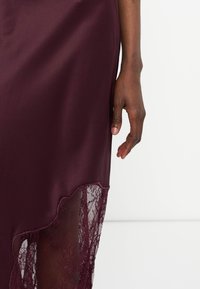 Main à la peau foncée à côté d'une jupe en satin bordeaux avec un détail en dentelle transparente près de l'ourlet sur un fond clair uni.