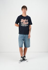 Tamno plava majica s bijelim i crvenim natpisom, uparena s svijetlim traper šortsvima. Crne visoke tenisice upotpunjuju ležerni outfit.