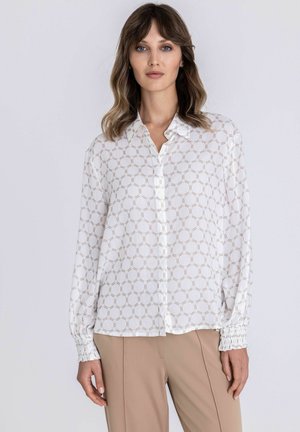 Woman wearing a white patterned button-up blouse with long sleeves and beige trousers, standing against a plain light background.