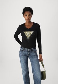 Svart langermet skjorte med v-hals, med en triangel-logo i gull og leopardmønster, utsmykket med sølvspiker. Kombinert med blå jeans.