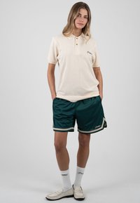 Femme debout, les mains dans les poches, portant un polo beige, un short de sport vert foncé, des chaussettes blanches et des mocassins beiges sur un fond uni.