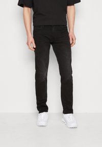 Jeans de mezclilla negra de corte slim, con cinco bolsillos, un sutil desgaste y cierre estándar de botón, combinados con zapatillas blancas.