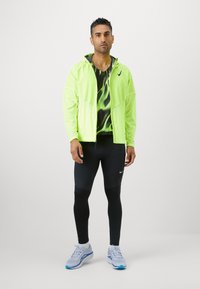 Giacca leggera verde neon con zip sopra una camicia nera a fantasia, abbinata a leggings neri e sneakers azzurre chiare con accenti.