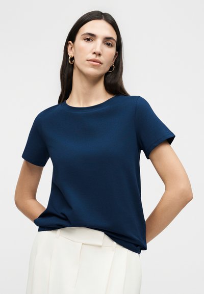 Γυναίκα με μακριά σκούρα μαλλιά, που φορά χρυσούς κρίκους στα αυτιά, μπλε ναυτικό τοπ με κοντά μανίκια και ψηλόμεσο λευκό παντελόνι, στέκεται με τα χέρια πίσω από την πλάτη.