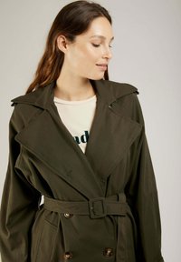 Trench-coat vert olive avec une taille ceinturée, des revers surdimensionnés, un devant croisé et des poches latérales, fabriqué en tissu lisse.
