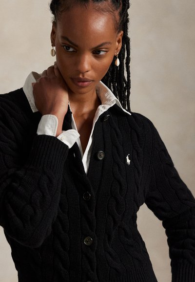 Nő fonott hajjal, kagylófülbevalót visel, fehér galléros ing és fekete, kötött mintás kardigán kis fehér logóval.