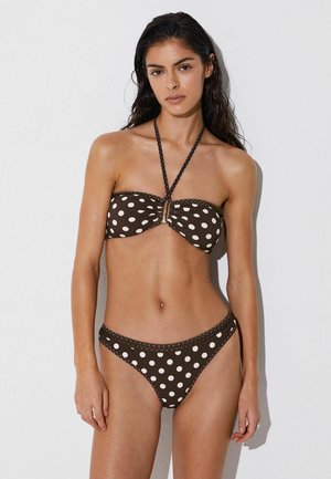 Giovane donna con capelli lunghi e scuri che indossa un bikini marrone con grandi pois bianchi, in piedi contro una parete bianca.