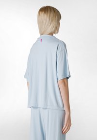 Camisa de manga corta azul claro en tela suave con un ajuste holgado. Presenta un pequeño detalle de logo rosa en el cuello trasero.