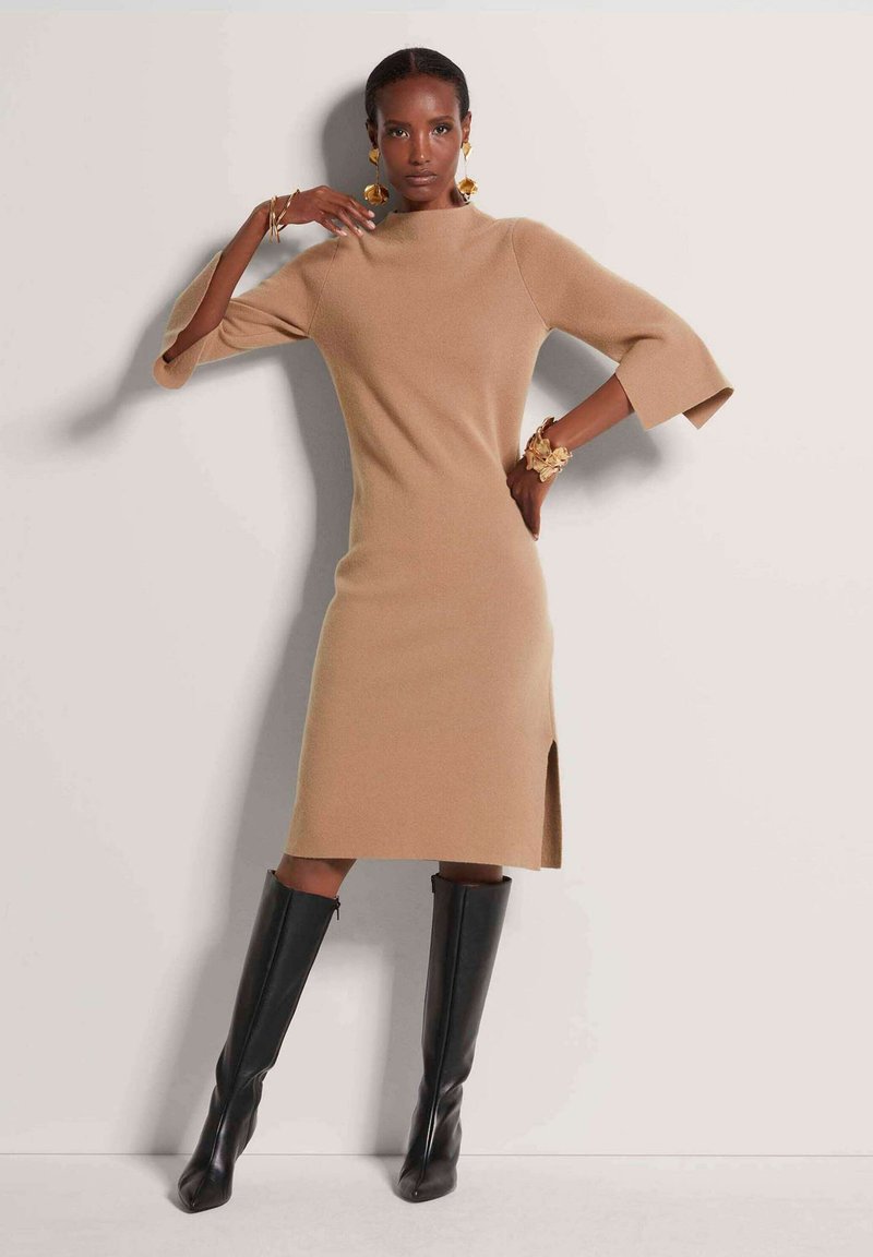Beiges Strickkleid mit Ellbogenlangen Ärmeln und einem seitlichen Schlitz, kombiniert mit hohen schwarzen Stiefeln. Glatte Textur, figurbetontes Design.