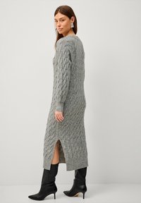Robe en tricot grise avec un motif en cable, à manches longues et fentes sur les côtés, associée à des bottes noires ajustées au genou.