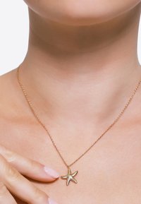 Gouden zeester hanger ketting op een delicate ketting, met gedetailleerde textuur, gedragen tegen een neutrale huidtint achtergrond.