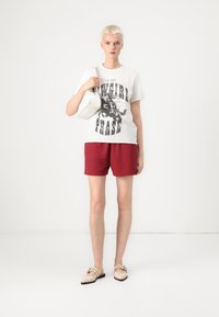 T-shirt blanc avec un texte graphique noir, short rouge et ballerines beiges. Tient un petit sac blanc. Le mannequin se tient sur un fond neutre.