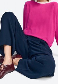 Henkilö istuu päällään leveät tummansiniset housut, kirkkaan vaaleanpunainen pitkähihainen paita, valkoinen t-paita, ja burgundin väriset nauhakengät valkoista taustaa vasten.