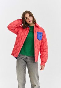 Žena v korálově růžové prošívané bundě s modrým kapsou a zeleném svetru pózuje s jednou rukou za hlavou na bílém pozadí.