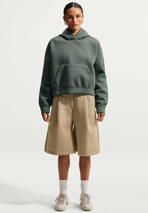 Kvinde stående iført en grøn hættetrøje, beige brede knælange shorts, hvide sokker og beige sneakers mod ensfarvet baggrund.