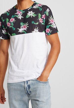 Mann trägt kurzärmeliges T-Shirt mit schwarzem Oberteil, das rosa und weiße Blumen zeigt, und weißem Unterteil, kombiniert mit hellblauen Jeans.