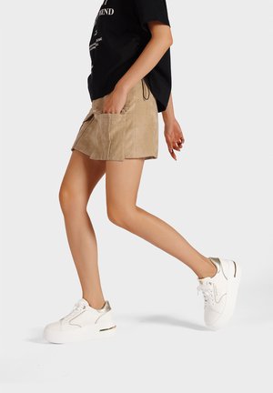 Gonna in suede beige con due tasche frontali, abbinata a sneakers bianche con dettagli dorati e suola spessa. Modella in cammino a metà passo.