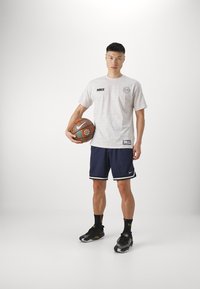 Camiseta blanca de Nike con patrón circular y logo negro; pantalones cortos navy con rayas blancas; zapatillas negras. Modelo sujetando un baloncesto.